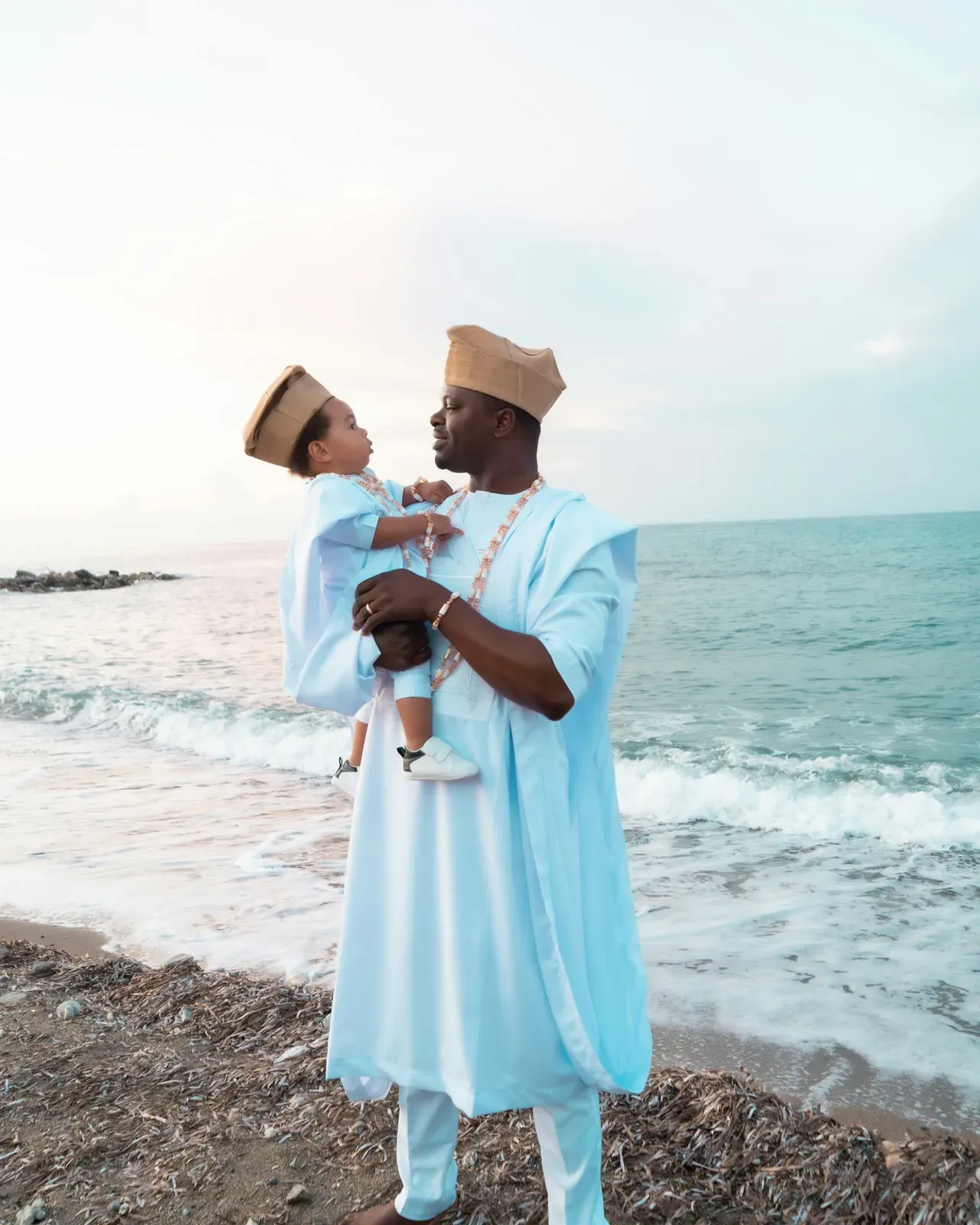 Man in a flowing pale blue-white agbada and matching cap holding his young son, also dressed in white with a cap, on a beach