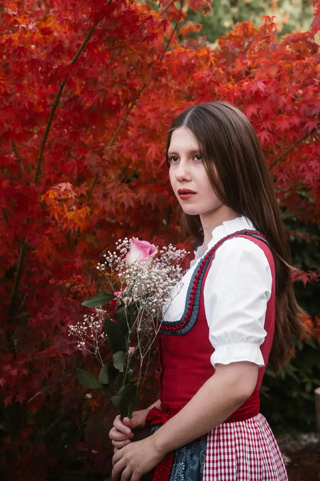 Young woman in a dirndl with a red bodice, white puff-sleeved blouse and red-and-white checked apron, holding a bouquet with a pink rose and baby's breath in front of red autumn leaves