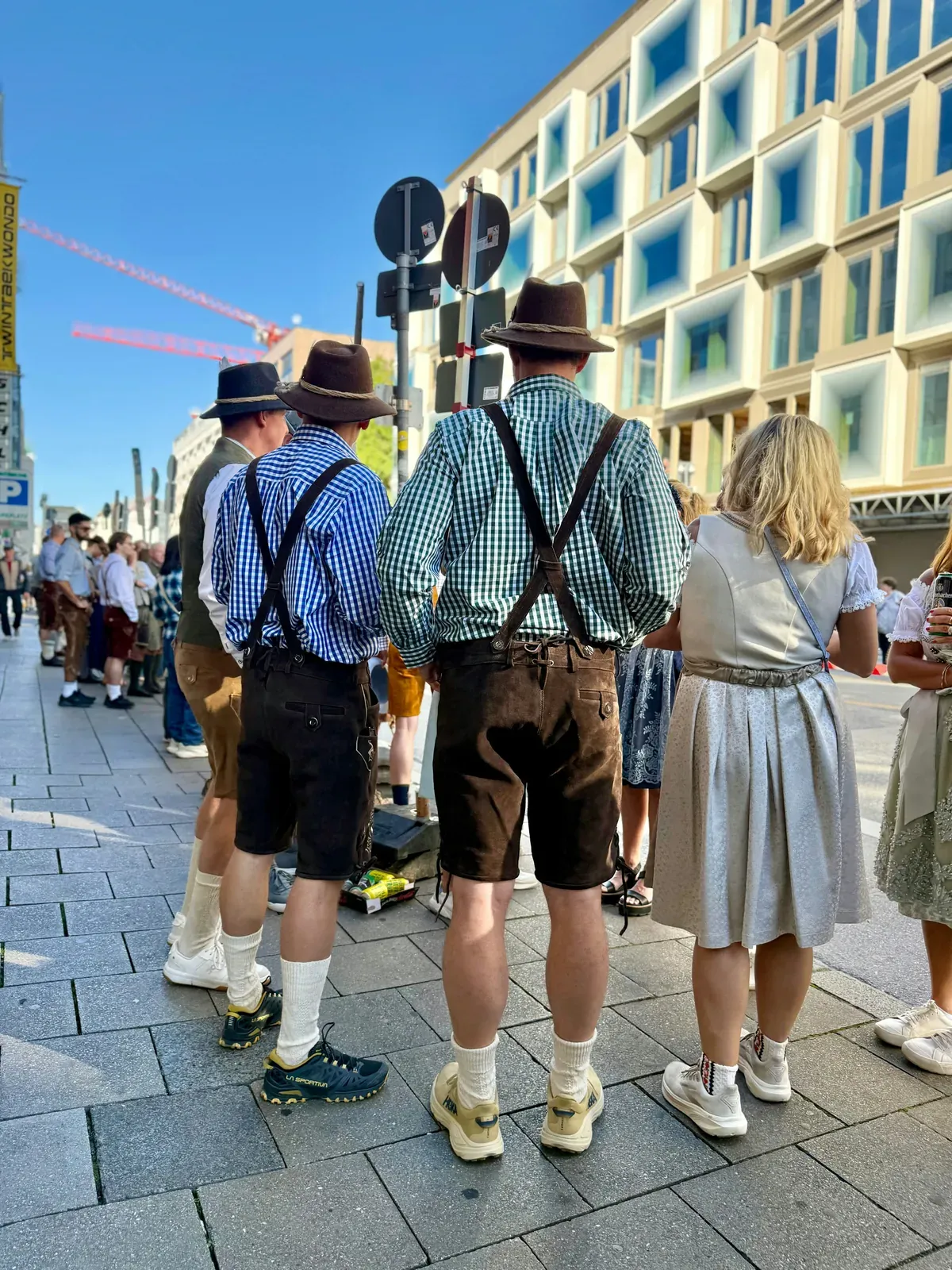 People seen from behind on a sunny sidewalk in Munich, men in leather lederhosen with suspenders, checked shirts and felt hats, and a woman in a light blue-grey dirndl