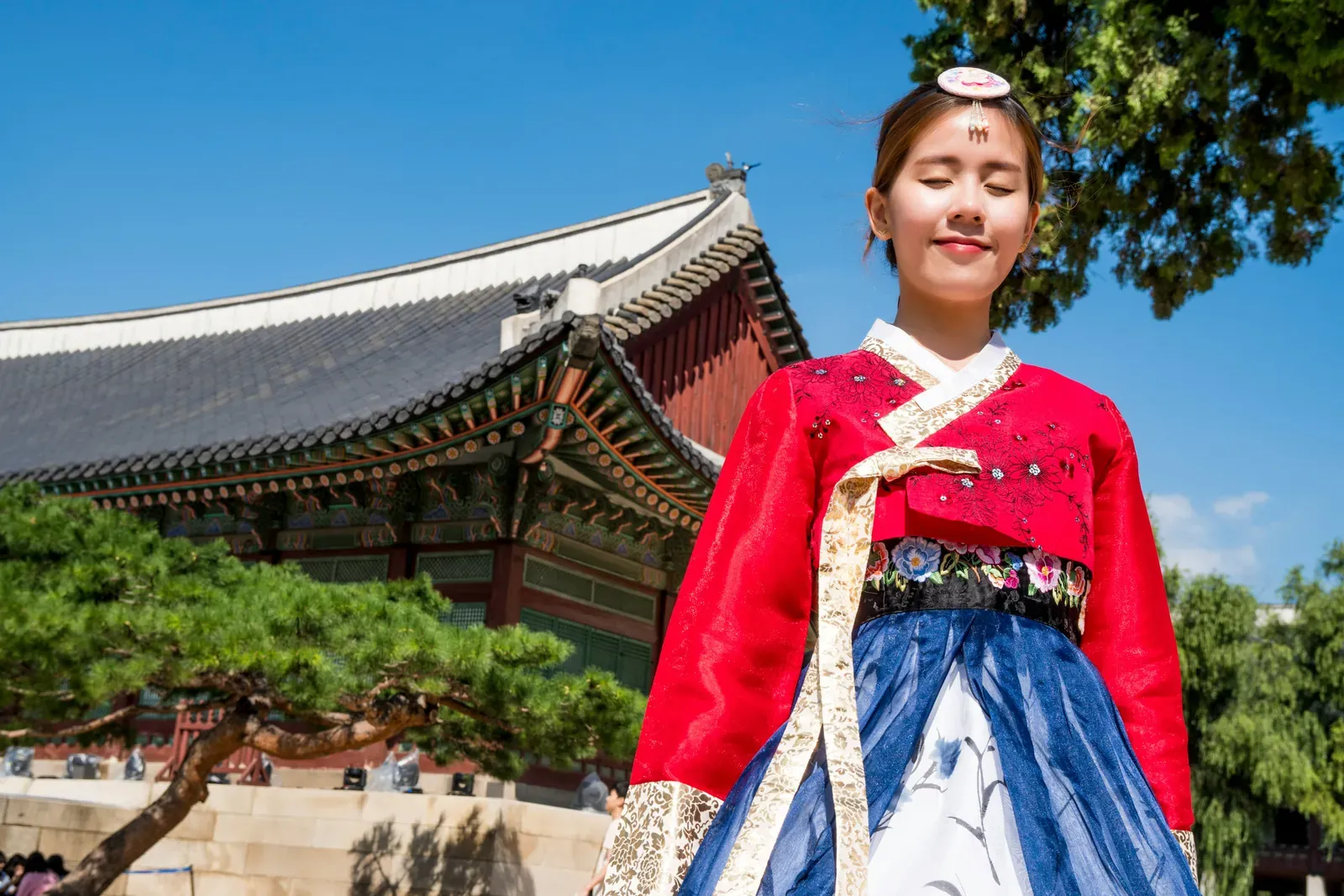 Smiling young woman in a red jeogori jacket with white collar trim and a blue chima skirt in front of a building at Gyeongbokgung Palace