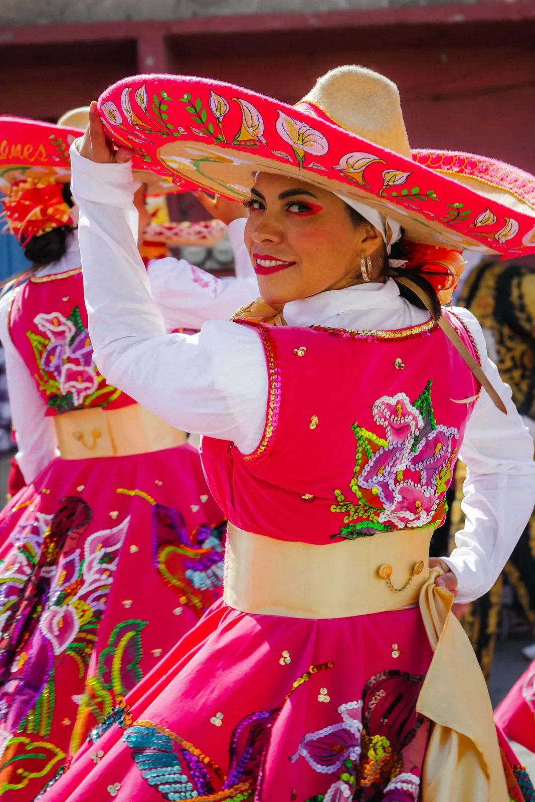 Smiling woman at a festive dance performance in a bright pink dress with large colourful floral embroidery, white sleeves and a yellow sash, lifting a wide red-and-white embroidered sombrero