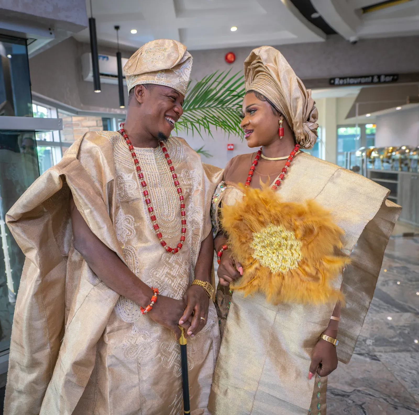 Bride and groom in matching gold attire at a traditional wedding in Nigeria, the bride in a large gele head tie, the groom in a flowing robe and cap