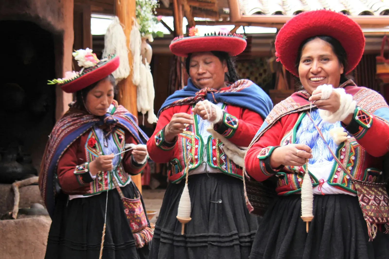 Three smiling women in red embroidered jackets, black skirts, woven shawls and flat round red hats, spinning yarn with drop spindles