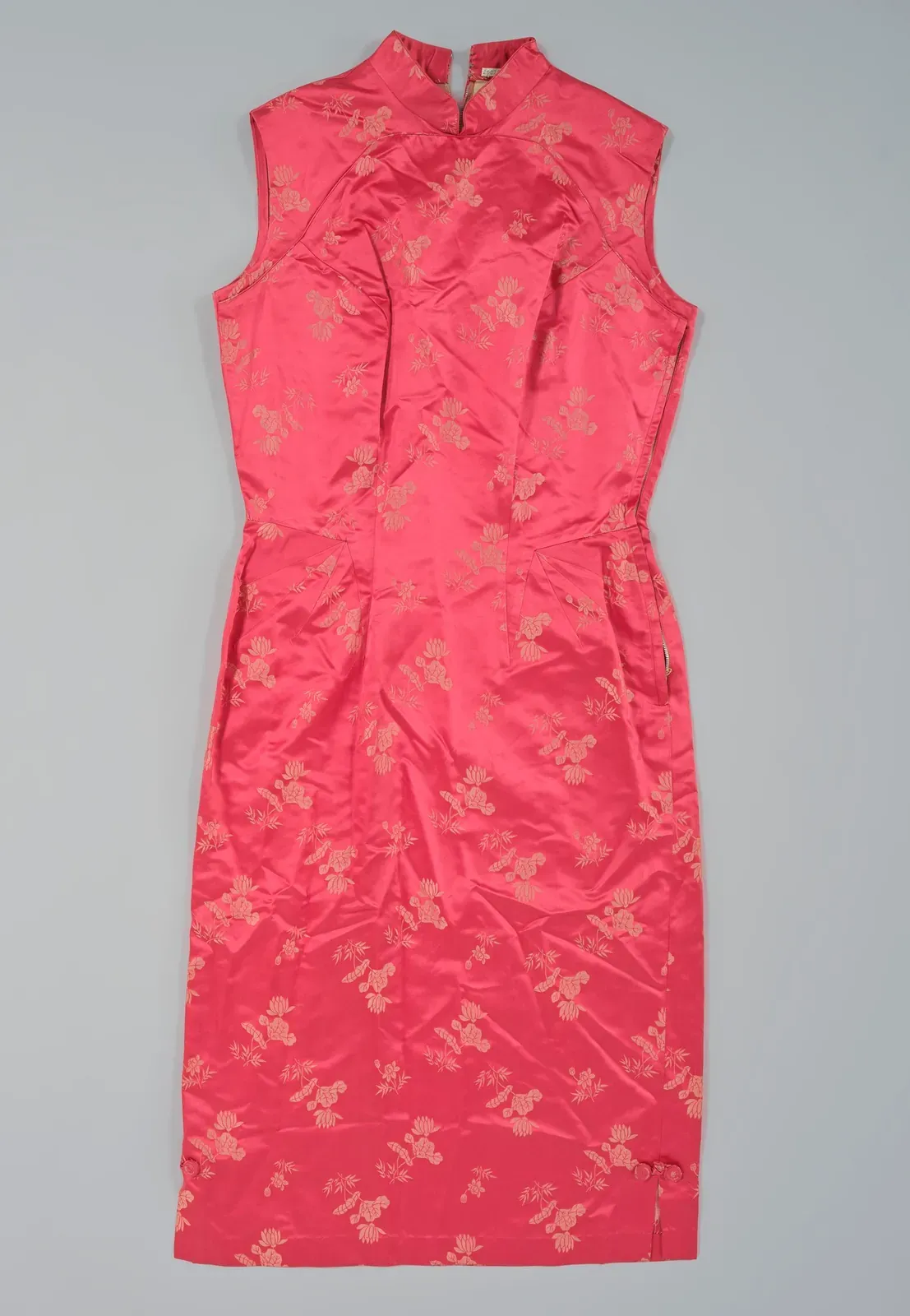 Fitted sleeveless coral-red qipao with a high mandarin collar and a small woven floral pattern, on a plain grey background